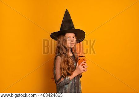 Little Witch Girl Holding Festive Glass With Polka Dots And Looking At Camera On A Yellow Background