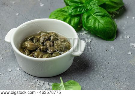 Capers. Marinated Or Pickled Canned Capers Fruit With Fresh Basil Leaves In A Small Bowl, On Gray Ba