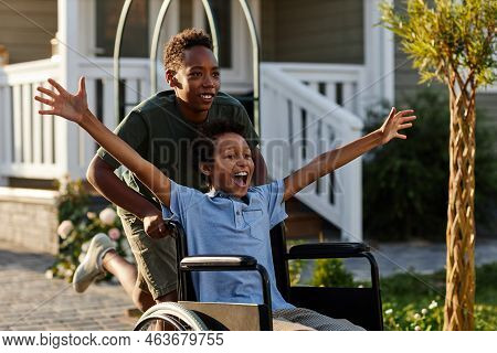 Portrait Of Black Teenage Boy Pushing Littlle Brother In Wheelchair While Having Fun Together Outdoo