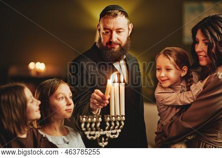 Portrait Of Orthodox Jewish Man Lighting Menorah Candle With Family During Hanukkah Celebration
