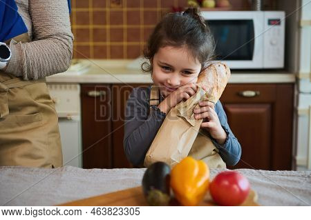 Adorable Child, Cute Baby Girl In A Beige Chefs Apron, Holds A Hot Baguette Of Freshly Baked Whole G