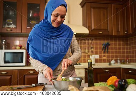 Beautiful Middle Eastern Woman Wearing A Blue Hijab, Using A Wooden Spoon, Mixing Ingredients And Fr