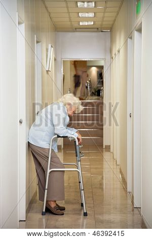 Full length of a tired elderly woman with walker standing in hospital passageway