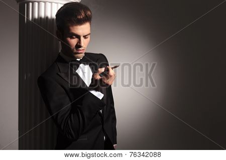 Attractive young business man leaning on a white column while holding a cigarette in his right hand, looking down.