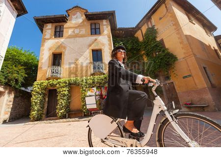 Girl with black elegant dress on a bicycle in the village square