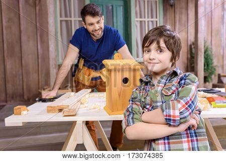 Happy father leaning at table with tools and birdhouse while looking at smiling boy with crossed arms