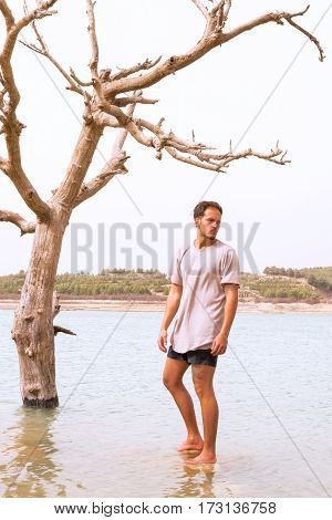 handsome young taned male model stood under a dying tree in a flooded lakeconcept of climate change. filter added blank sky in background for copy space