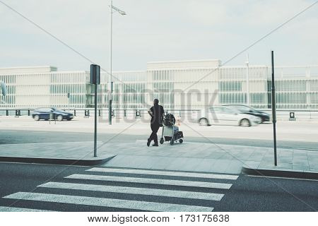 Rear view of man with luggage cart standing near airport zebra traffic cross way silhouette of experienced male with several suitcases waiting for taxi outdoors near airport terminal in Barcelona