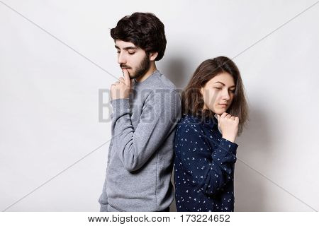Unhappy couple standing back to back holding their hands on chins looking down having thoughtful expressions. Bearded guy and young brunette having some misunderstanding between each other