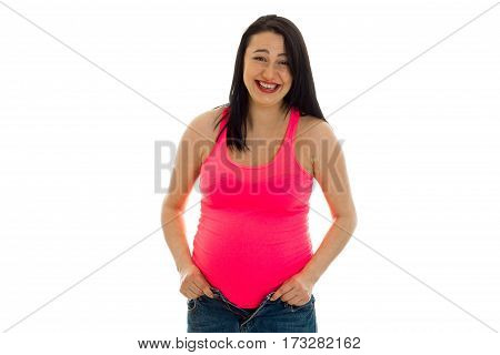 Young pregnant woman in a bright pink tank top is laughing because her blue jeans don't fit anymore isolated on a white background.