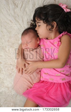 Portrait of a 3 1/2 year old sister kissing her 9 day old baby sister. Shot from overhead on a white flokati rug.