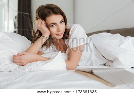 Image of young cheerful woman dressed in white t-shirt lies in bed at home indoors. Looking at camera.