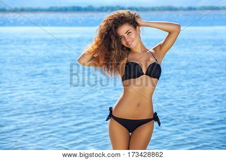 Young, Attractive, Smiling Slender Girl With Curly Hair In A Black Bathing Suit On The Beach.