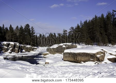 Wide view of a snowy country landscape with a small stream of rapids running through rock faces on either side and a background of green fir trees at Pabineau Falls near Bathurst, New Brunswick, on a bright sunny day with blue skies and clouds in February
