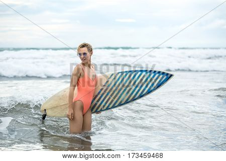 Delightful blonde girl with short hairstyle stands sideways in the water on the beach on the background of the sea and the cloudy sky. She wears orange swimsuit with sunglasses and holds a surfboard.