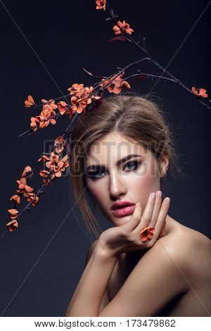 Beautiful young woman with make-up and loose hairdo holding artificial sakura branch with orange flowers. Beauty shot on dark background. Copy space.