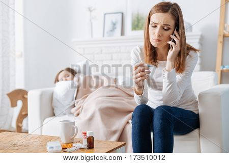 What we can do. Cheerless caring troubled woman calling a doctor for her ill daughter who is lying on the sofa in the background