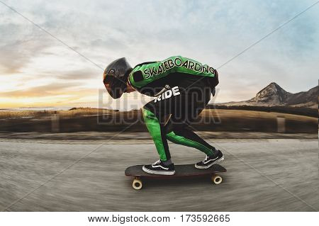 A man in a fullface helmet and a leather suit in a special rack rides a longboard on afsaltu sunset in the background mountains and beautiful sky at high speed.