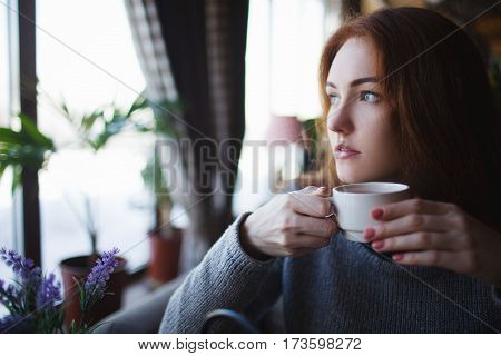 Red-haired girl in sweater drinking coffee and looking out the window