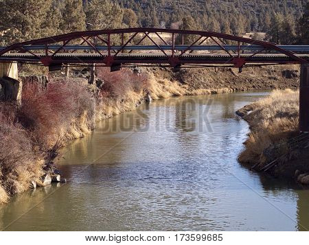 An old bridge spans the Crooked River in Central Oregon with bushes and rocks on the banks.