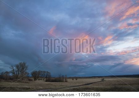 Sunset.Dirt road through the field. Remote leafless trees.Red sunlight falls on a dark cluster of clouds unusual shape.DecemberUkraine