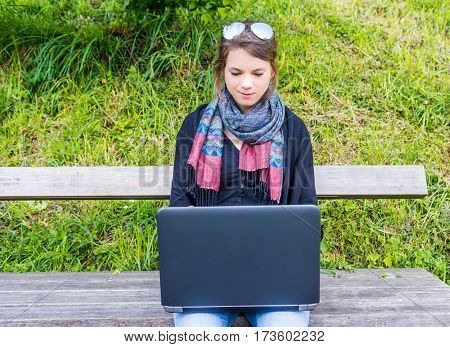 Young woman using laptop at a riverbank. Milleanial working in a park.