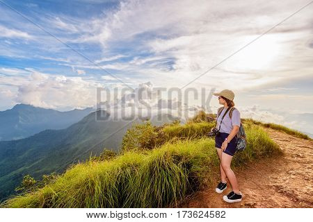 Hiker asian teen girl happy with camera backpack cap and eyeglass looking beautiful landscape nature of mountain range and colourful sky at sunset on Phu Chi Fa Forest Park Chiang Rai Thailand