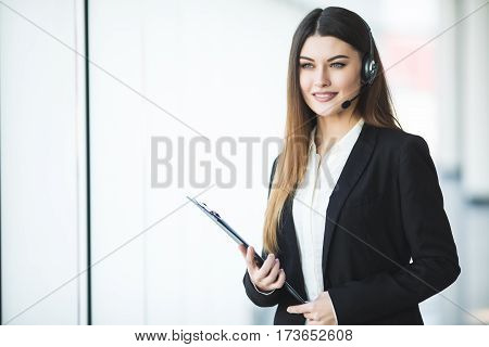Photo of beautiful young call center operator standing in office. Woman with headphones looking at camera and smiling