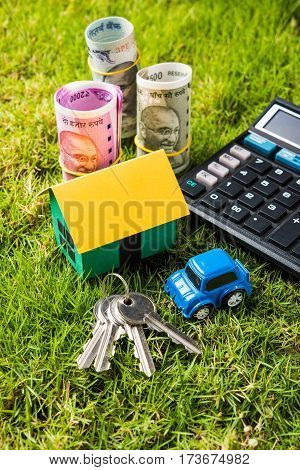 indian currency notes of rupees 100, 500 and 2000 in roll or stack with small home model, keys, toy car and calculator with blank board over green grass - home buying and finance concept in India