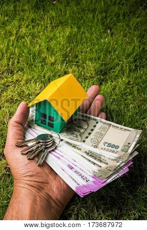 hand holding new indian currency notes or rupees 500 and 2000 and house model over green grass - indian real estate and finance concept
