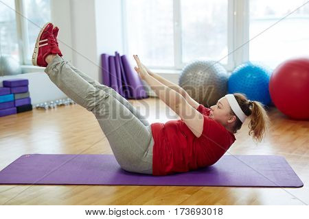 Side view portrait of determined obese woman working out in fitness studio: performing toe tap straight leg sit ups on yoga mat