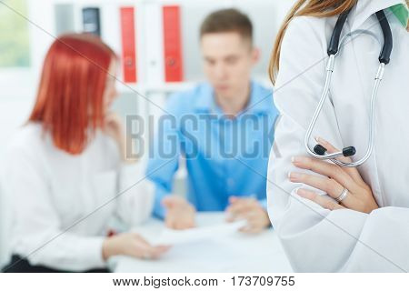 Female medicine doctors's hands crossed on her chest closeup with family couple on the background. Medical help or insurance concept. Doctor is waiting for patient to examine. Physician reception concept