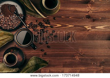 Hot coffee in black cup and several turkish pots cezve with beans dry leaves with copy space on brown old wooden board background top view. Rustic style.