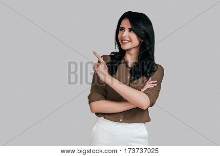 Look over there! Beautiful young woman in smart casual wear keeping arms crossed and looking away with smile while standing against grey background