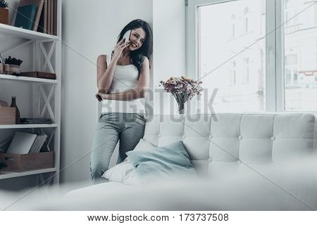 So good to hear from you! Attractive young woman talking on the mobile phone and smiling while leaning on the wall at home