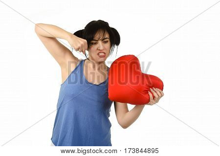 young attractive and beautiful woman punching in rage spiteful and resentful a red heart shape pillow upset and angry isolated background in love pain and romantic relationship broken concept