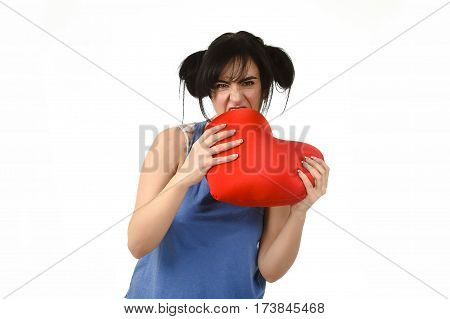 young attractive and beautiful woman biting in rage spiteful and resentful a red heart shape pillow upset and angry isolated background in love pain and romantic relationship broken concept