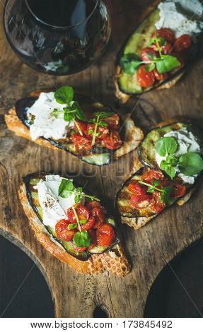 Wine and snack set. Brushetta with grilled eggplant, cherry tomatoes, garlic, cream cheese, arugula and glass of red wine on wooden board over dark background, selective focus. Slow food concept
