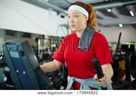 Cute overweight girl with red hair working out using machines in gym, dreaming to be thinner