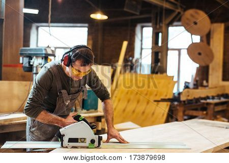 Man in protective eyeglasses and headphones cutting wooden plank on workbench