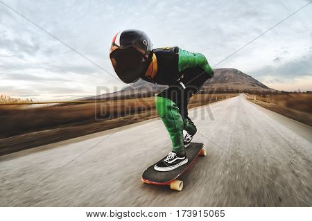 A young man in a fullface helmet and a leather suit in a special rack rides a longboard on afsaltu sunset in the background mountains and beautiful sky at high speed.