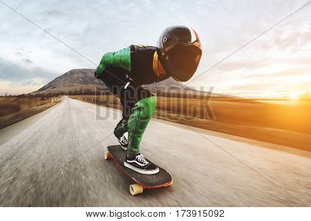 A young man in a fullface helmet and a leather suit in a special rack rides a longboard on afsaltu sunset in the background mountains and beautiful sky at high speed.