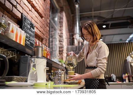 small business, people and service concept - happy woman or barmaid pouring smoothie from blender to cup at vegan cafe