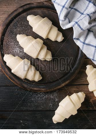 Raw unbaked croissant on wooden chopping board with kitchen towel over old dark wood background. Top view