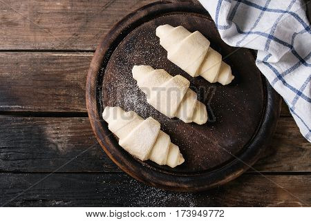 Raw unbaked croissant on wooden chopping board with kitchen towel over old dark wood background. Top view