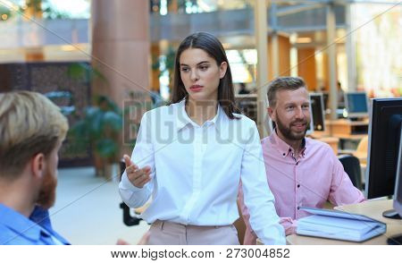 Group Of Young People In Casual Wear Sitting At The Office Desk And Discussing Something While Looki