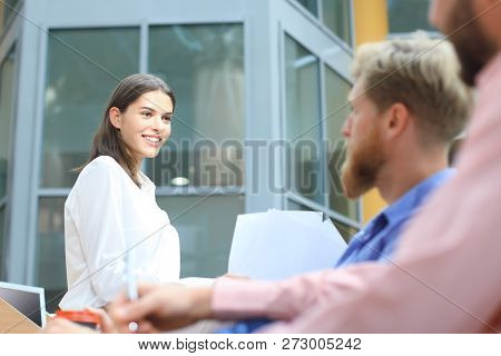 Group Of Young People In Casual Wear Sitting At The Office Desk And Discussing Something While Looki