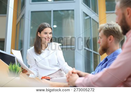 Group Of Young People In Casual Wear Sitting At The Office Desk And Discussing Something While Looki