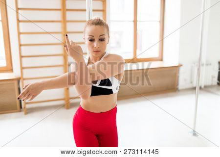 Young fit woman stretching left arm while bending the other one during poledance workout or warm-up