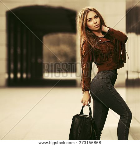 Young fashion woman with handbag walking on city street Stylish female model wearing brown leather fringe suede jacket and dark blue jeans